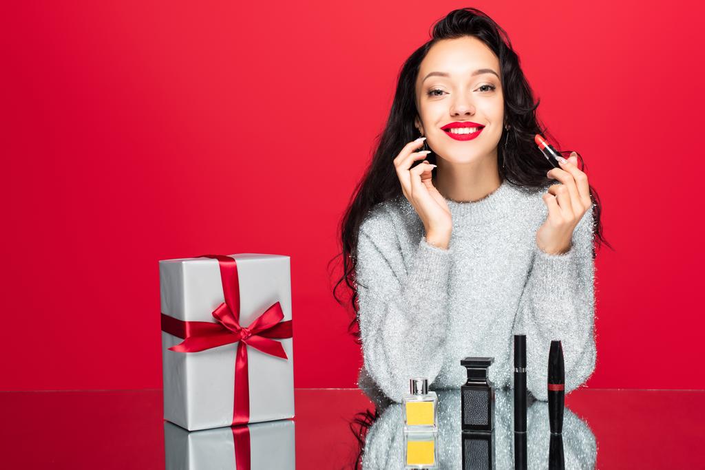 весела жінка в светрі тримає помаду біля декоративної косметики, пляшка з парфумами і загорнута в подарунок ізольована на червоному
 - Фото, зображення