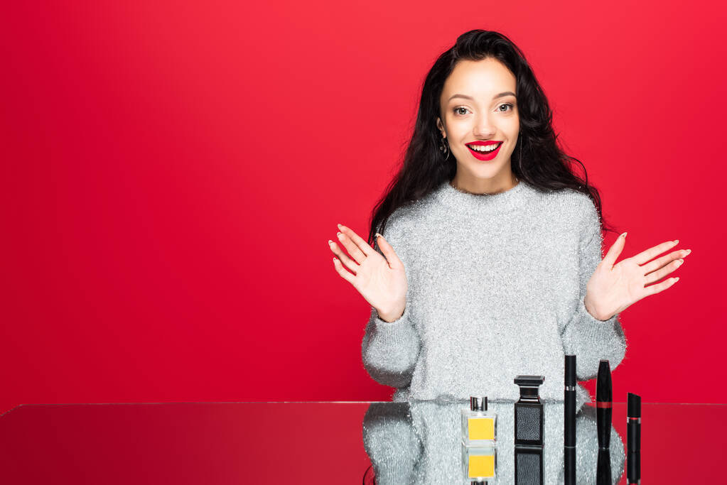 усміхнена жінка в светрі жестикулює біля декоративної косметики і пляшок з парфумами ізольовані на червоному
 - Фото, зображення