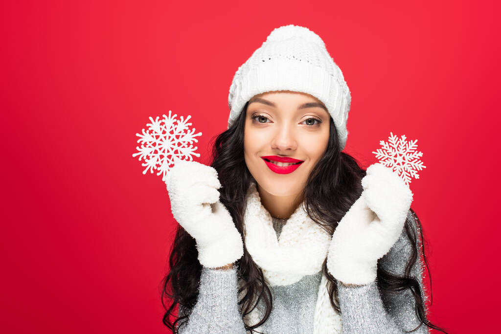 pleased woman in winter outfit holding decorative snowflakes isolated on red - Photo, Image