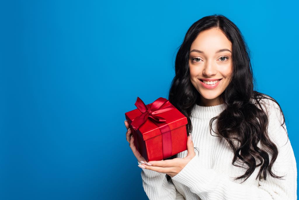 cheerful brunette woman in knitted sweater holding christmas present isolated on blue  - Photo, Image