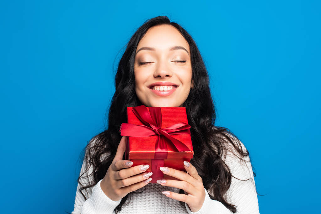 glückliche brünette Frau mit geschlossenen Augen hält Weihnachtsgeschenk isoliert auf blau  - Foto, Bild
