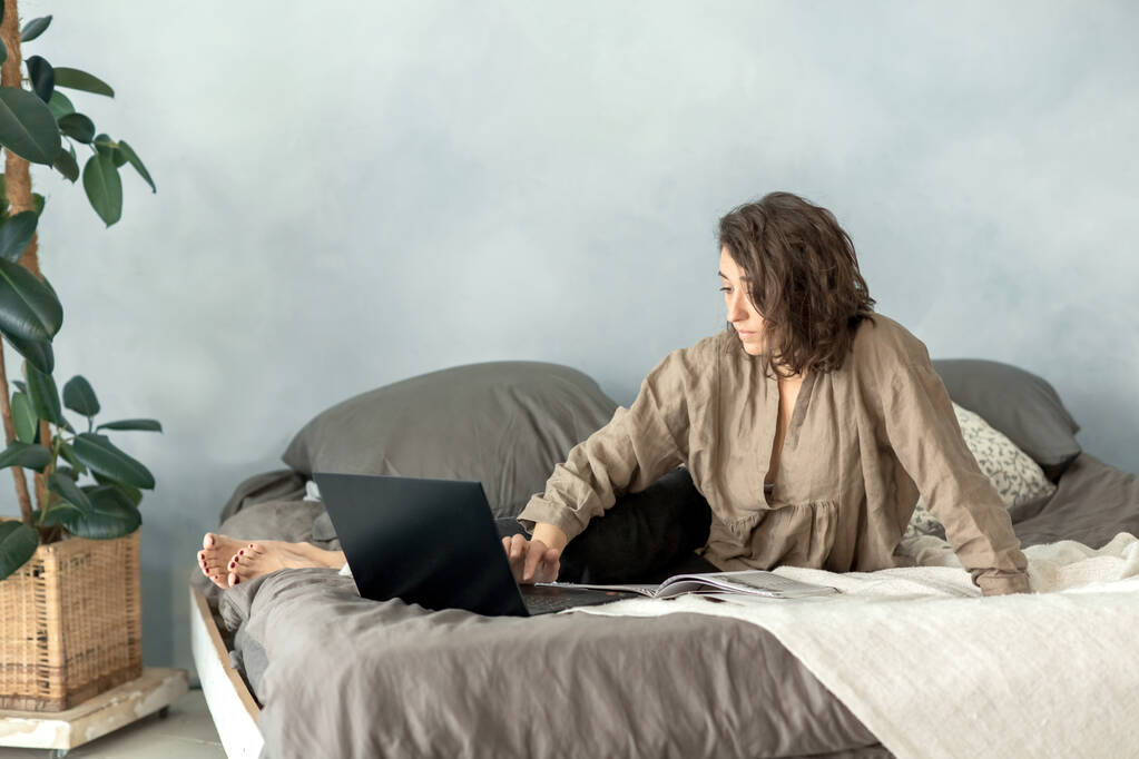 Egy fiatal barna nő laptopot használ a hálószobájában, miközben a kanapén ül. Laptopot használ, hogy filmet nézzen, beszélgessen a barátaival, könyvet olvasson, és e-mailt nézzen. - Fotó, kép