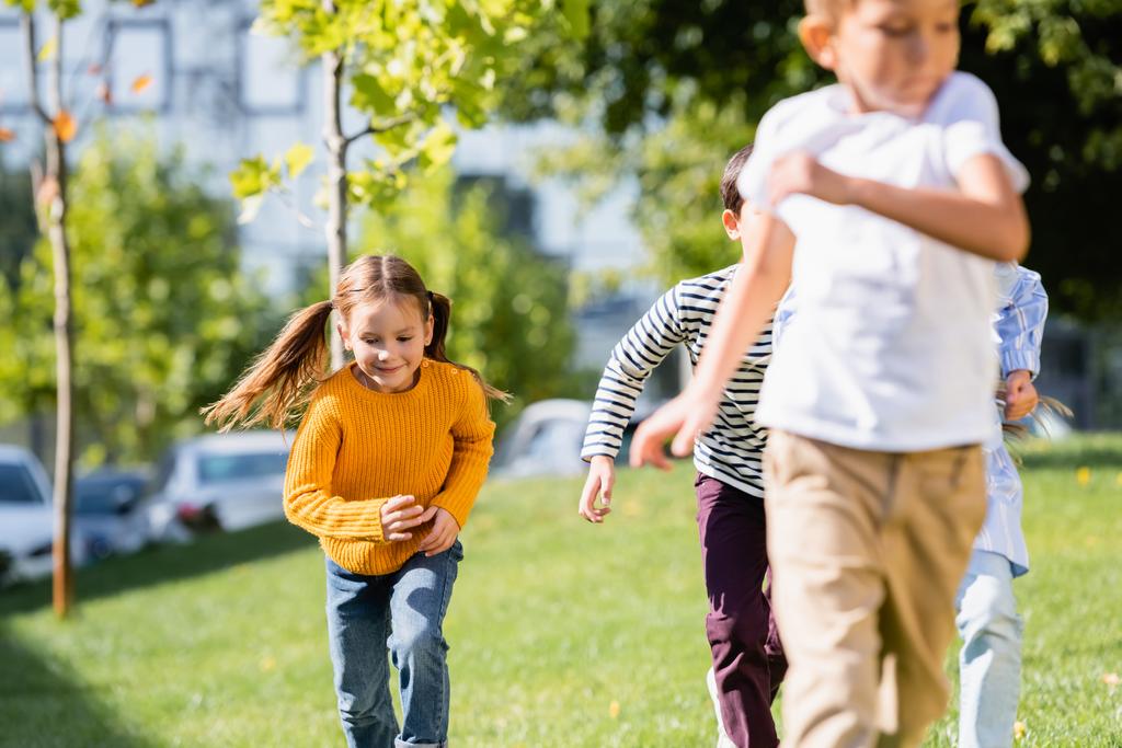 Χαμογελαστή κοπέλα τρέχει κοντά σε φίλους σε θολή πρώτο πλάνο στο πάρκο  - Φωτογραφία, εικόνα