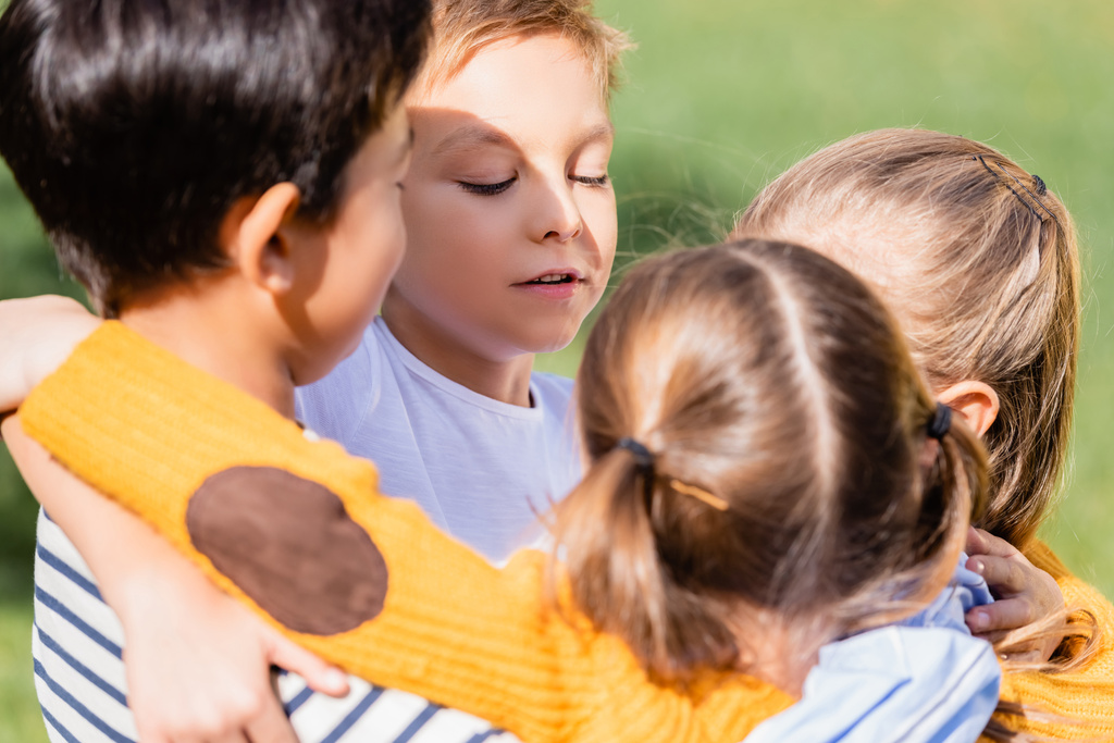 Boy hugging multiethnic friends on blurred foreground outdoors  - Photo, Image
