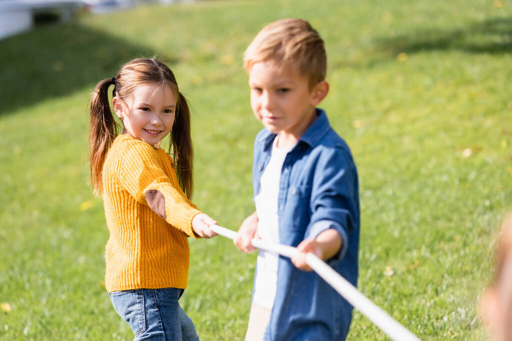 Bambino positivo giocare tiro alla fune vicino amico in primo piano sfocato nel parco  - Foto, immagini