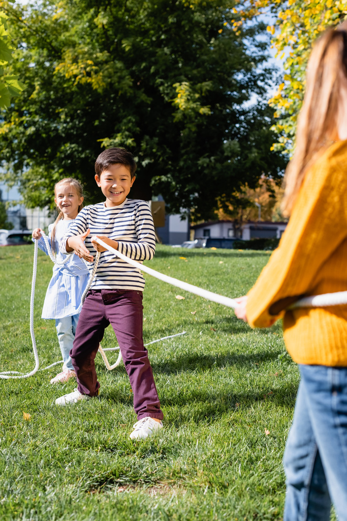 sonriendo asiático chico tirando cuerda mientras jugando tirón de la guerra con amigos en borrosa primer plano en parque  - Foto, imagen