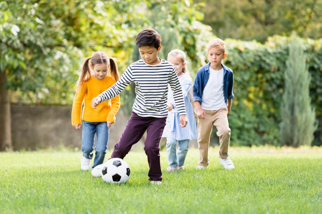 Азиатский мальчик играет в футбол рядом с друзьями на размытом фоне в парке  - Фото, изображение