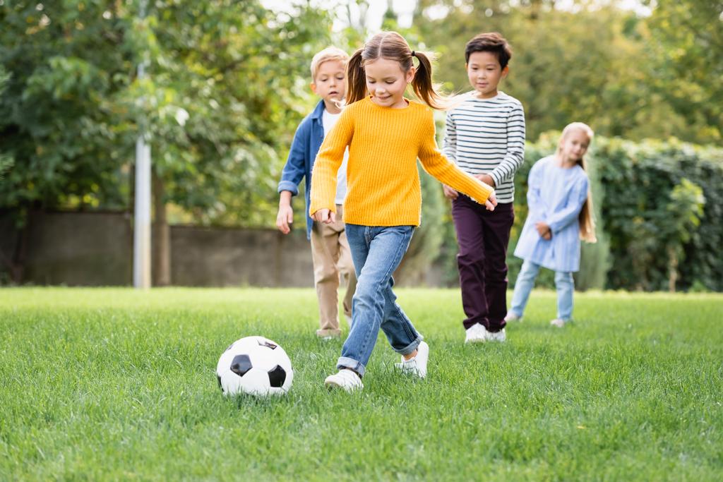 Улыбающаяся девушка играет в футбол рядом с многонациональными друзьями на размытом фоне в парке  - Фото, изображение