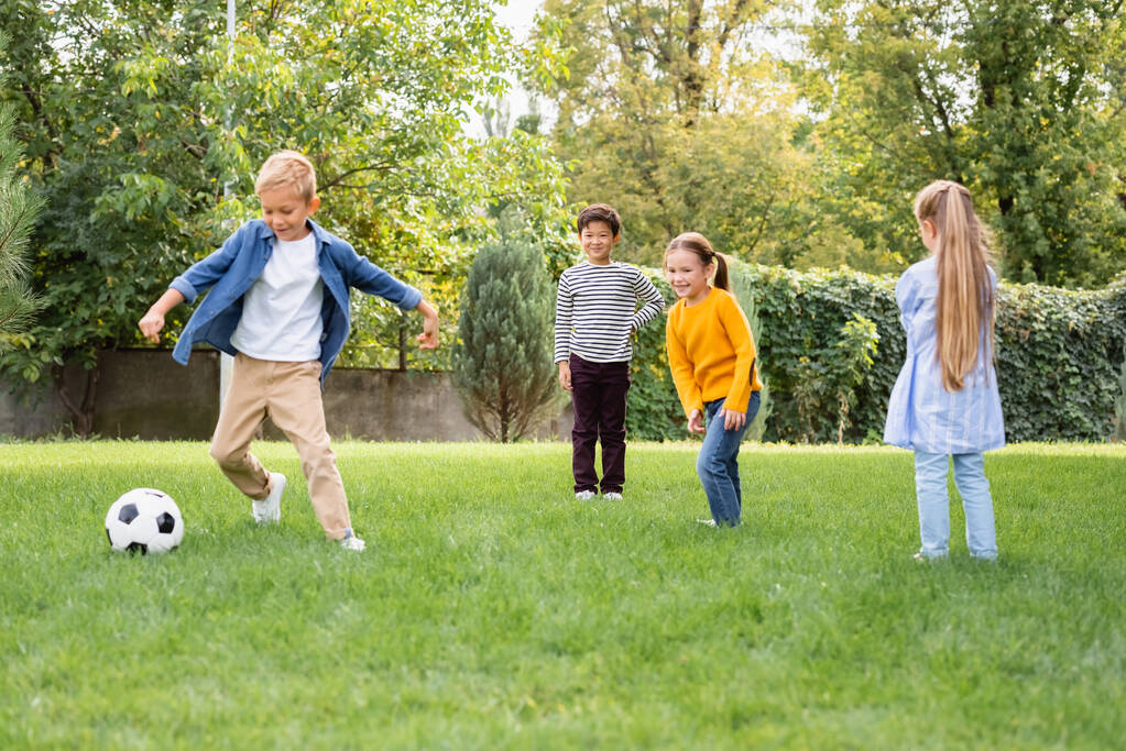 Positiivinen monikansallinen lapset seisoo lähellä ystävä pelaa jalkapalloa puistossa  - Valokuva, kuva