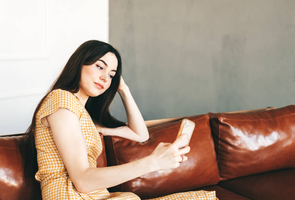 Młoda kaukaska kobieta korzystająca ze smartfona podczas odpoczynku na stylowej sofie w domu. Wysokiej jakości zdjęcie - Zdjęcie, obraz
