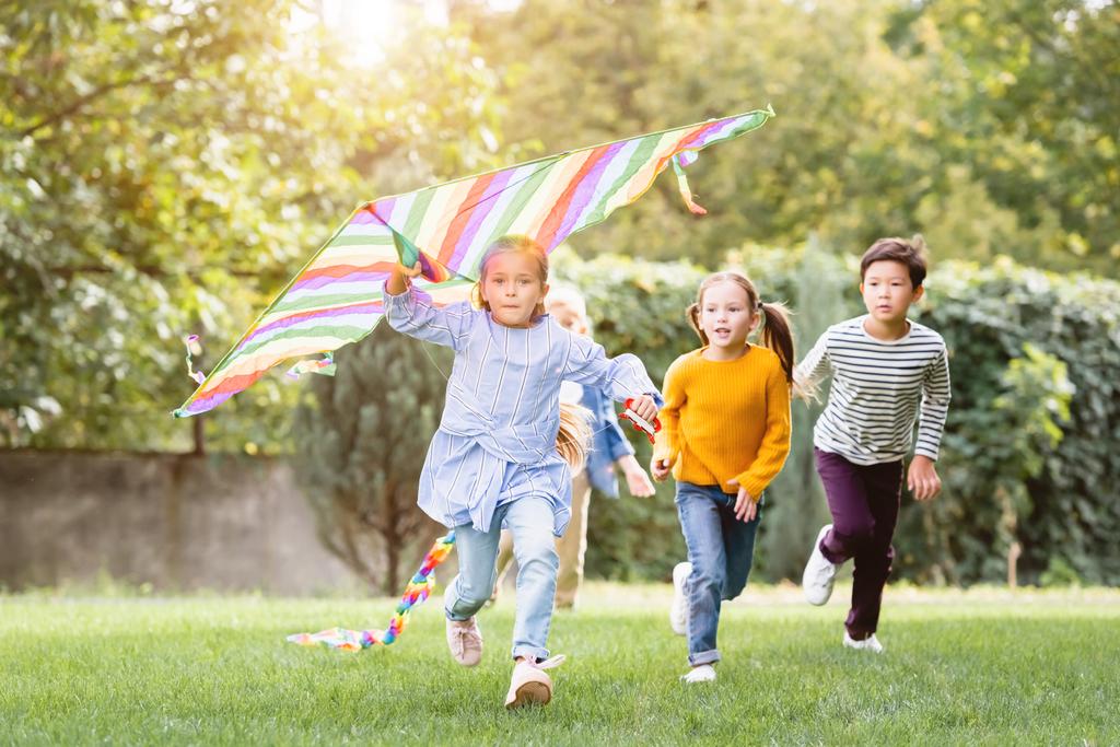 Girl holding flying kite while running near multiethnic friends in park  - Photo, Image