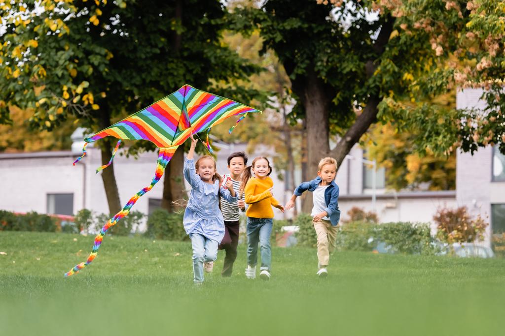 Χαμογελώντας πολυεθνικά παιδιά τρέχουν στο γκαζόν, ενώ παίζει με ιπτάμενο χαρταετό στο πάρκο  - Φωτογραφία, εικόνα