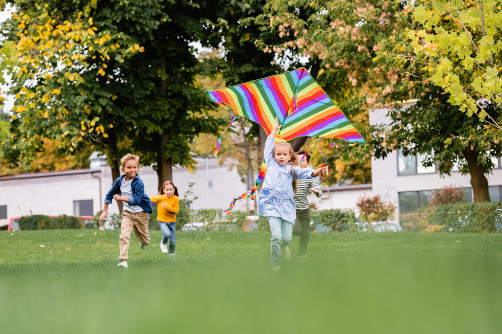 Chica sosteniendo colorido volando cometa mientras juega con amigos multiétnicos en fondo borroso en el parque  - Foto, Imagen
