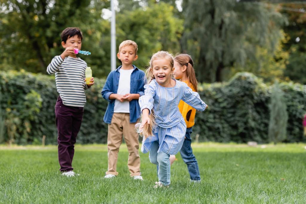Lächelndes Mädchen läuft auf Rasen neben multiethnischen Freunden mit Seifenblasen auf verschwommenem Hintergrund  - Foto, Bild