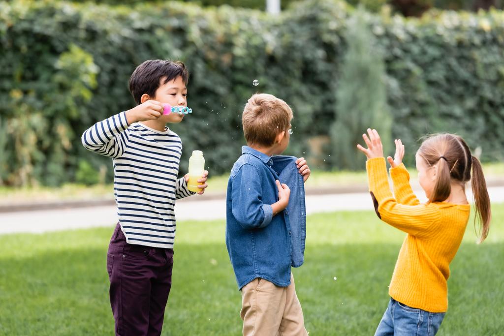 Asian schoolboy blowing soap bubbles near friends in park  - Photo, Image