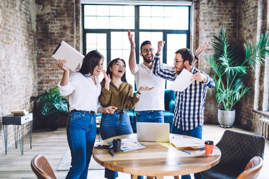 Группа счастливых многорасовых коллег в повседневной одежде улыбается и стоит вокруг стола в офисе, прыгая с поднятыми руками - Фото, изображение