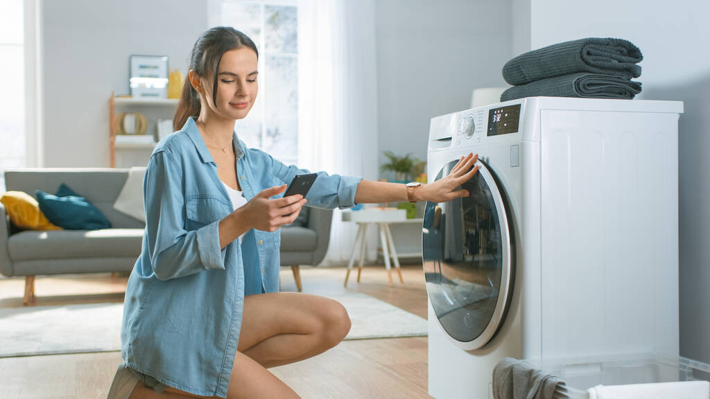 Die schöne junge Frau sitzt auf ihren Knien neben der Waschmaschine. Sie lud die Waschmaschine mit schmutziger Wäsche, während sie ihr Smartphone benutzte. Schuss im Wohnzimmer mit modernem Interieur. - Foto, Bild