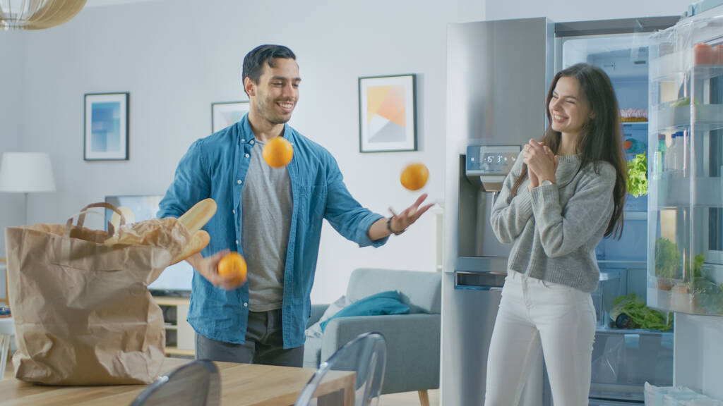 Beautiful Young Couple Have Fun in the Kitchen. Man is Juggling with Oranges. Girl is Clapping, Cheering for Him and They Laugh. Room Has a Modern Tech Fridge with Fresh Groceries. - Photo, Image