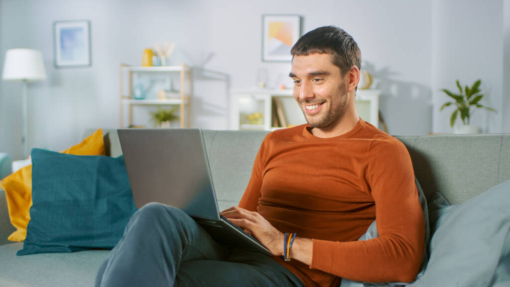 Homem bonito usa computador portátil enquanto sentado no sofá em casa. Homem trabalhando, navegando através da Internet a partir de sua acolhedora sala de estar. - Foto, Imagem