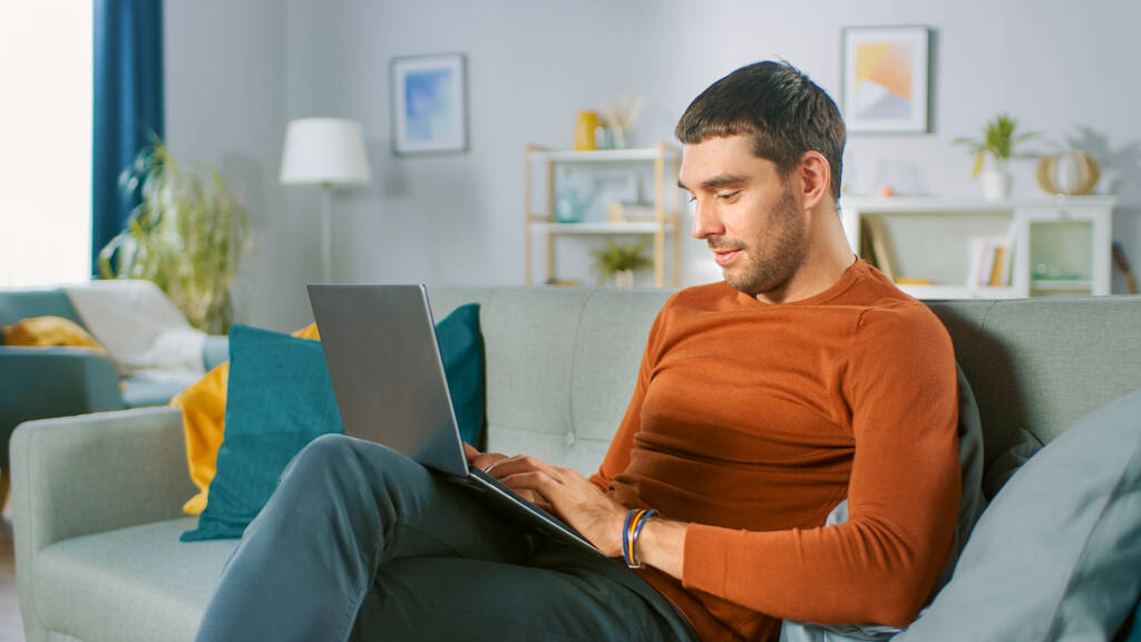 Fešák používá notebook, zatímco sedí na pohovce doma. Muž pracující, procházení internetu ze svého útulného obývacího pokoje. - Fotografie, Obrázek