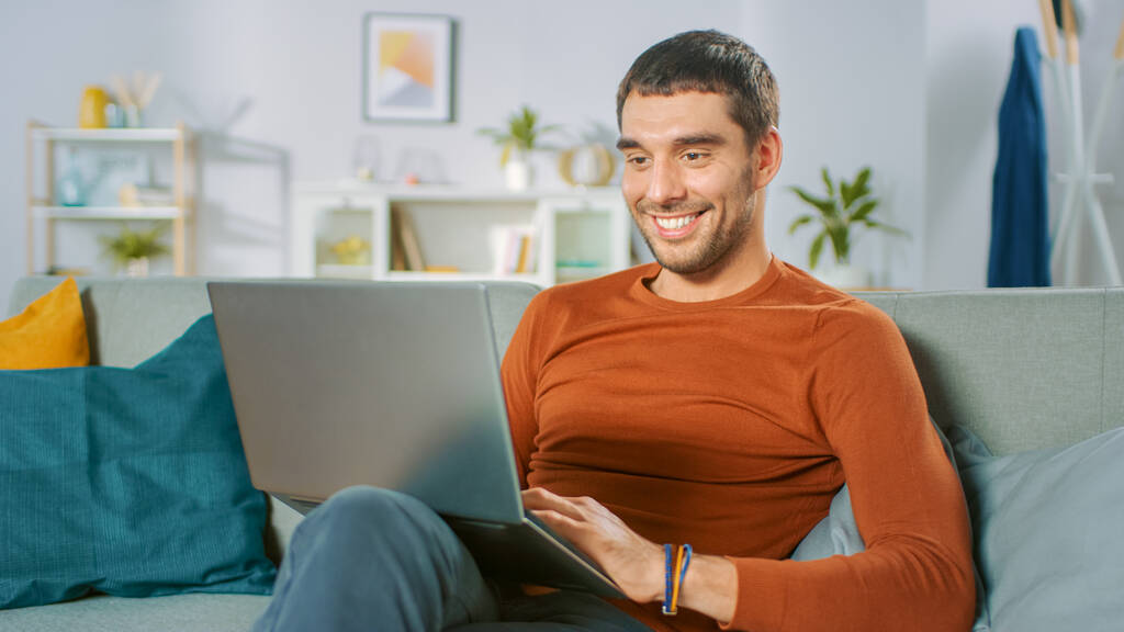 Hombre guapo utiliza ordenador portátil mientras está sentado en el sofá en casa. El hombre trabajando, navegando a través de Internet desde su acogedora sala de estar. - Foto, imagen