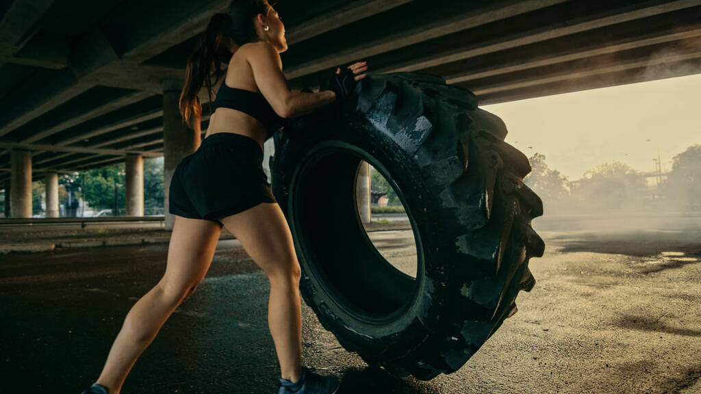 Schöne selbstbewusste Fitness-Mädchen in schwarzem athletischen Top macht Übungen auf der Straße. Schuppen kippen einen großen schweren Reifen in einer städtischen Umgebung. - Foto, Bild