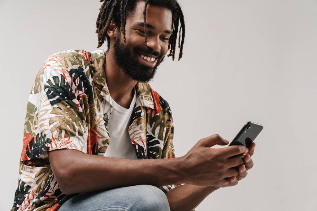 Obrázek pozitivního usmívajícího se afrického chlapa izolované přes šedé stěny pozadí pomocí mobilního telefonu - Fotografie, Obrázek
