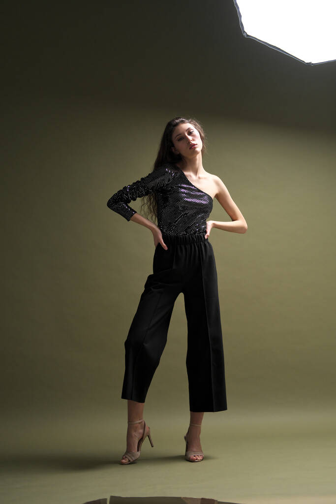A beautiful young tall girl with long black hair poses in a shiny blouse and black culottes on a green background. Buty and fashion photo shoot. - Photo, Image