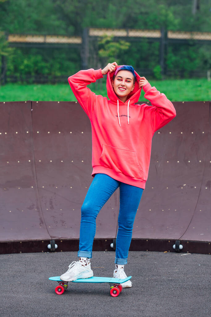 Портрет модной молодой девушки в розовой толстовке и джинсах, стоящей рядом со скейтбордом со своим голубым пластиковым скейтбордом. - Фото, изображение