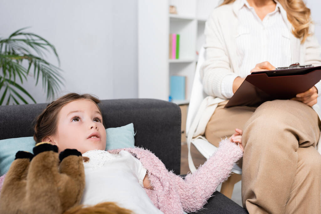 niña acostada en el sofá con juguete mientras le cuenta el problema al psicólogo - Foto, imagen
