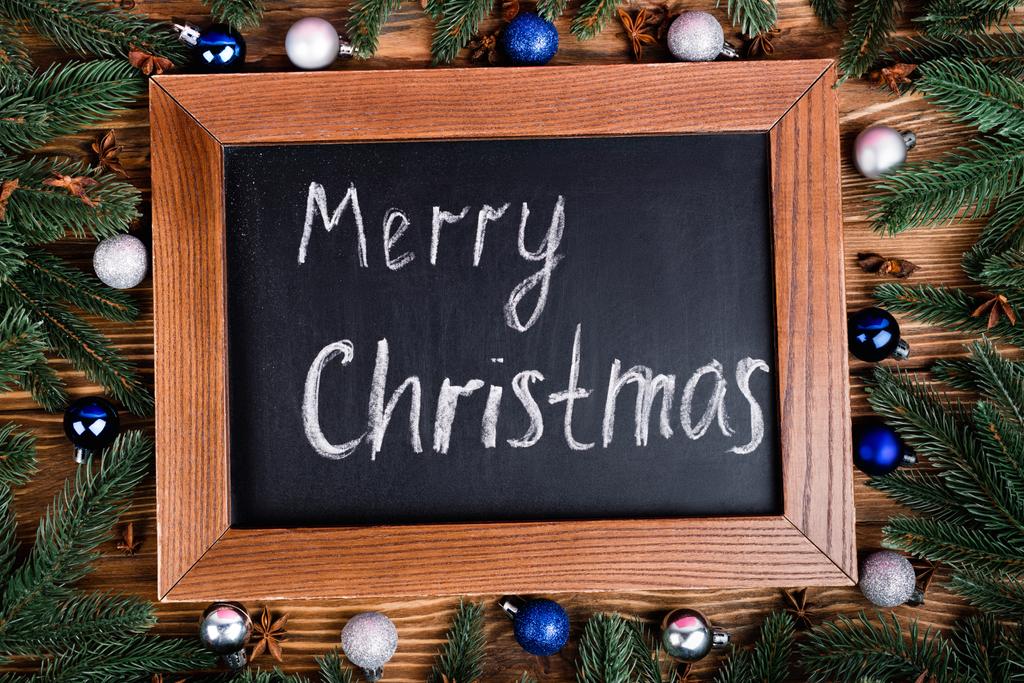 Bovenaanzicht van krijtbord met vrolijke kerstbelettering, dennentakken, kerstballen en anijssterren op bruine houten ondergrond - Foto, afbeelding