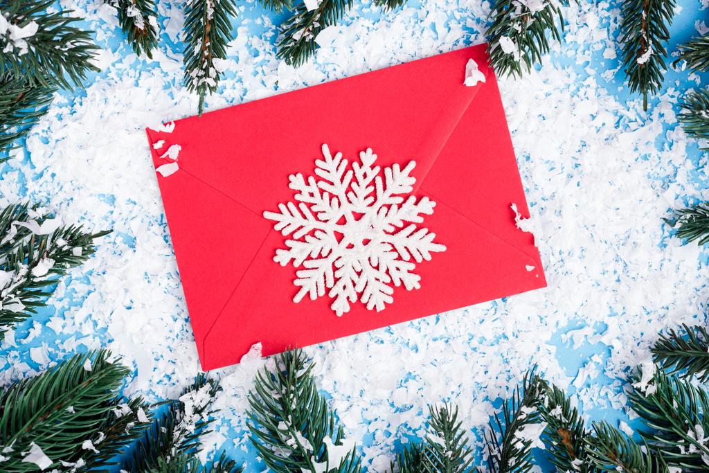 Vue de dessus du flocon de neige décoratif sur enveloppe rouge près des branches de pin et de la neige artificielle sur fond bleu - Photo, image