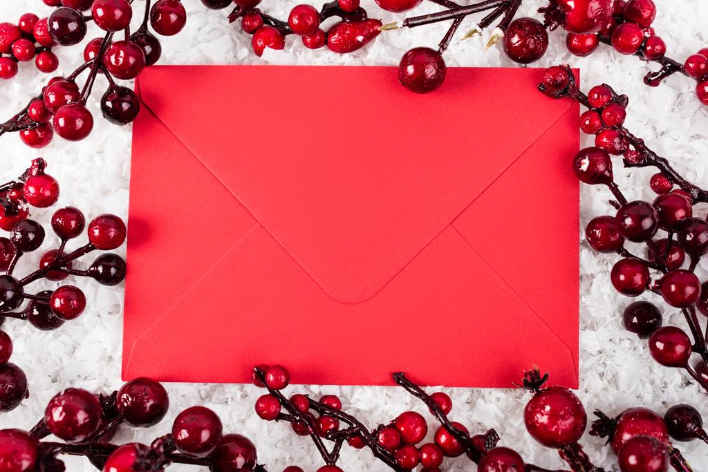 Вид зверху на червоний конверт біля гілок зі штучними ягодами на білому фактурному фоні
 - Фото, зображення