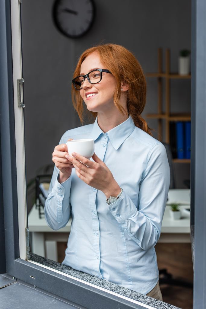Szczęśliwa ruda bizneswoman z filiżanką kawy odwracając wzrok, stojąc przy oknie z zamazanym biurem na tle - Zdjęcie, obraz