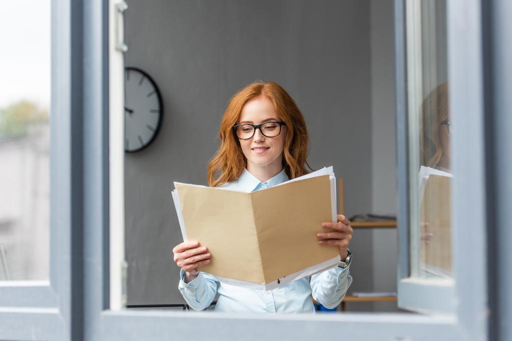 Улыбающаяся деловая женщина смотрит на папку с бумажными простынями в офисе с размытым окном на переднем плане - Фото, изображение