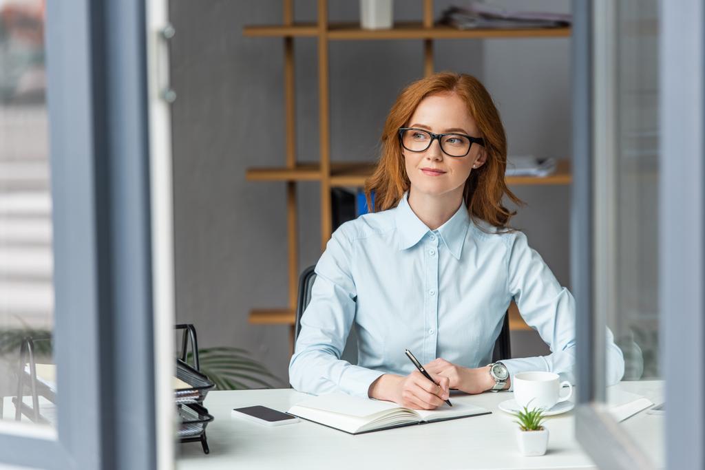 Позитивная деловая женщина, пишущая в блокноте, глядя в сторону на рабочее место с размытым окном на переднем плане - Фото, изображение