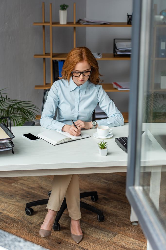 Longitud completa de la mujer de negocios escribiendo en el cuaderno, mientras está sentado en el lugar de trabajo con ventana borrosa en primer plano - Foto, imagen