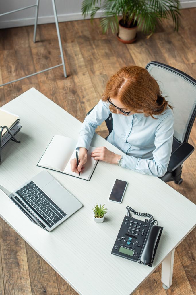 Overhead άποψη κοκκινομάλλα επιχειρηματίας γραφή στο σημειωματάριο, ενώ κάθεται στο χώρο εργασίας με συσκευές - Φωτογραφία, εικόνα