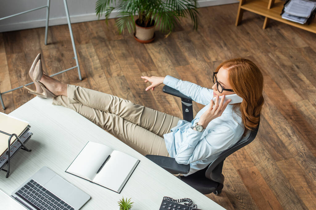 Vysoký úhel pohledu zrzka podnikatelka se zkříženýma nohama na stole mluvit na mobilním telefonu v kanceláři - Fotografie, Obrázek