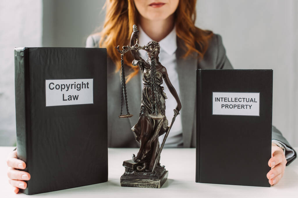 Ausgeschnittene Ansicht eines Rechtsanwalts, der Bücher mit Schriftzug über geistiges Eigentum und Urheberrecht in der Nähe der Figur auf dem Tisch vor verschwommenem Hintergrund hält - Foto, Bild