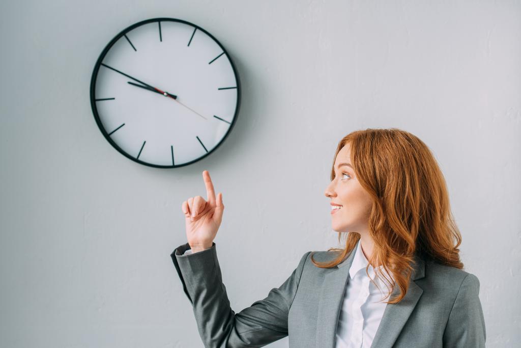Smiling redhead businesswoman pointing with finger and looking at wall clock on grey - Photo, Image