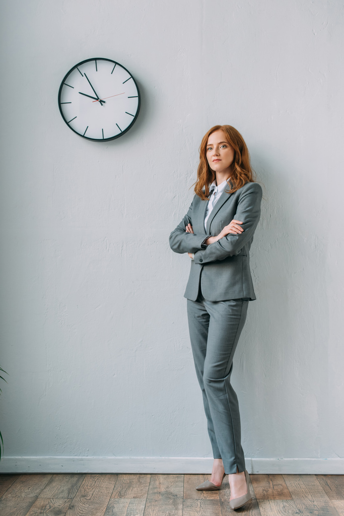 Повна довжина впевненої бізнес-леді з схрещеними руками, що стоять біля настінного годинника в офісі
 - Фото, зображення