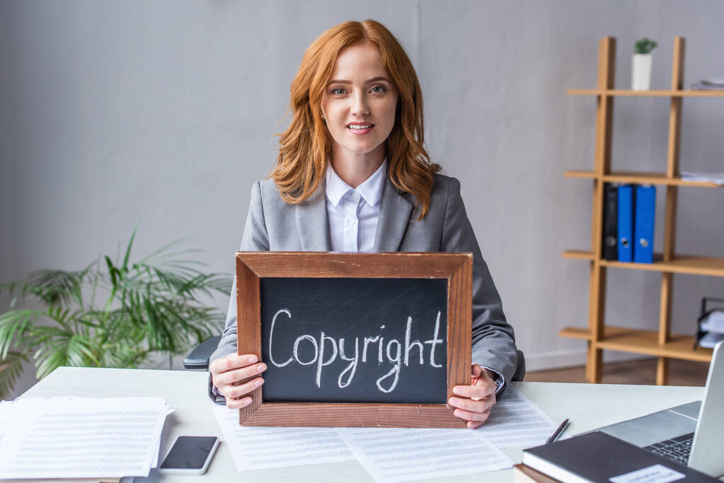 Lächelnder Anwalt zeigt Kreidetafel mit Urheberrechtsaufschrift, während er am Arbeitsplatz mit Dokumenten auf verschwommenem Hintergrund sitzt - Foto, Bild