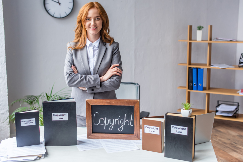 Uśmiechnięty prawnik ze skrzyżowanymi ramionami stojący przy tablicy z napisem autorskim w pobliżu książek na stole ze stosem dokumentów - Zdjęcie, obraz