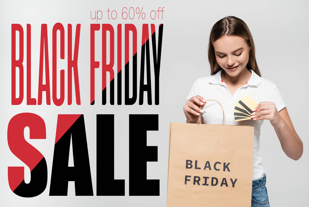 joyful woman holding credit cards and looking at shopping bag near black friday sale lettering on grey - Photo, Image