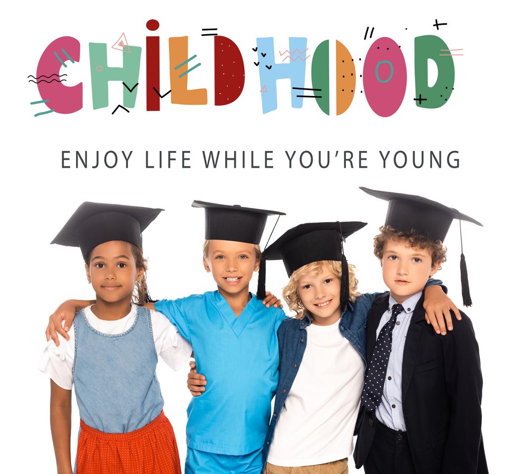 πολυπολιτισμικά παιδιά σε καπέλα αποφοίτησης ντυμένα με κοστούμια διαφόρων επαγγελμάτων κοντά στην παιδική ηλικία, απολαμβάνουν τη ζωή, ενώ είστε νέοι γράμματα σε λευκό  - Φωτογραφία, εικόνα