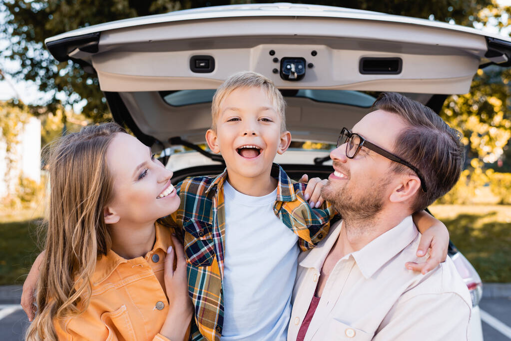 Sonrientes padres mirando al niño cerca de auto sobre fondo borroso al aire libre  - Foto, Imagen