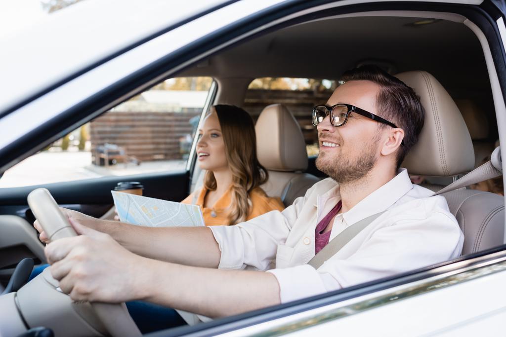 Sonriente hombre conduciendo coche cerca de la esposa con el mapa y el café para ir sobre fondo borroso  - Foto, Imagen