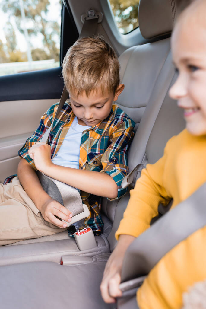 Niño bloqueando el cinturón de seguridad cerca de la hermana sonriente en primer plano borroso en el coche  - Foto, Imagen