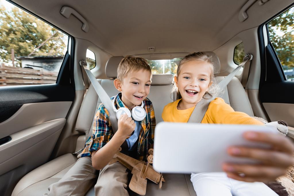 Enfants souriants avec jouets et écouteurs prenant selfie sur smartphone au premier plan flou dans la voiture  - Photo, image